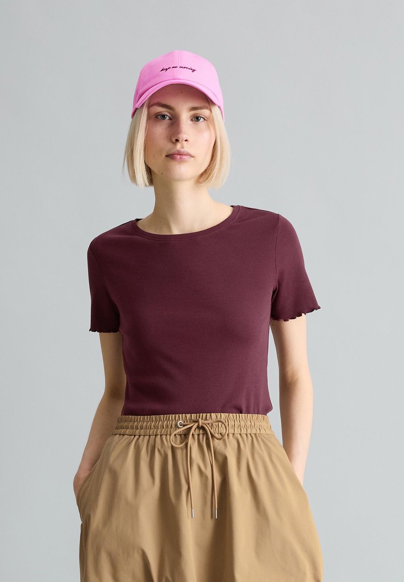 Giovane donna con taglio Bob biondo che indossa un cappellino rosa, una maglietta bordeaux a maniche corte e pantaloni beige con coulisse su uno sfondo grigio neutro.