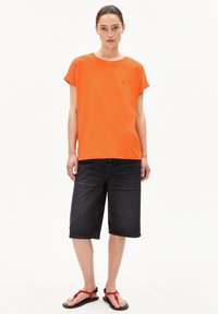 Oranges Baumwoll-T-Shirt mit hochgerollten Ärmeln, kombiniert mit dunklen Denim-Shorts. Schwarze Sandalen mit roten Akzenten runden das Outfit ab.