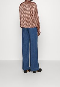 Kvinde iført bredbenede blå denimjeans, mønstret langærmet skjorte og sorte platåsko, der står foran en hvid baggrund.