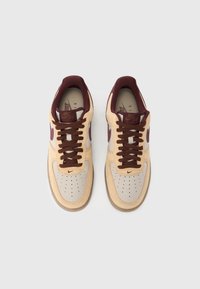 Beige ja burgundia värvi Nike Air Force 1 tossud pruunide paeltega, pealtvaade heleda halli taustal.