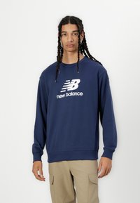 Tumši zils kokvilnas džemperis ar apaļu kakla izgriezumu, garām piedurknēm un balto "new balance" logo uz krūtīm. Iekļautas ribotas aproces un apakšmalas.