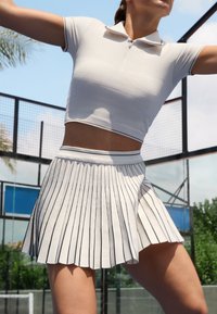 Top corto bianco a costine con colletto polo e zip, abbinato a una gonna bianca plissettata con strisce nere verticali.