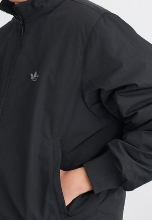 Personne portant une veste noire Adidas avec un col zippé, la main gauche dans la poche latérale, et le logo Adidas visible sur la poitrine.