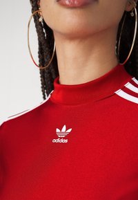 Κόκκινη αθλητική μπλούζα με ψηλό λαιμό, λευκό σχέδιο τριών λωρίδων και λογότυπο Adidas μπροστά. Το υλικό φαίνεται λείο και εφαρμοστό.