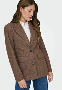 Bruine blazer met textuur, voorzien van een notch-kraag, twee voorzakken en twee zwarte knopen. Draagt over een wit shirt en in combinatie met donkere jeans.