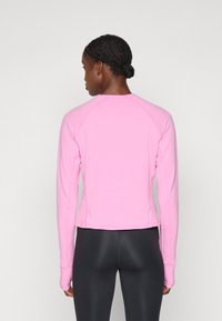 Maglia sportiva a maniche lunghe di un leggero rosa pallido con texture a coste, maniche raglan e design aderente, abbinata a leggings neri.