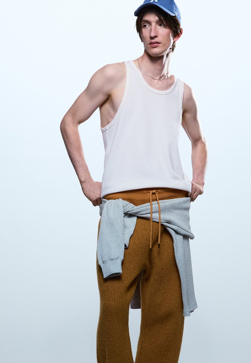 Νεαρός άνδρας που φοράει λευκό φανελάκι, μουσταρδί πλεκτό παντελόνι, μπλε καπέλο και γκρι φούτερ δεμένο γύρω από τη μέση, κοιτάζοντας πλαγίως.