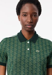 Grön pikétröja med geometriskt mönster, två knappar vid kragen och kontrasterande mörkgrön kant. Lacoste-logotyp på bröstet.