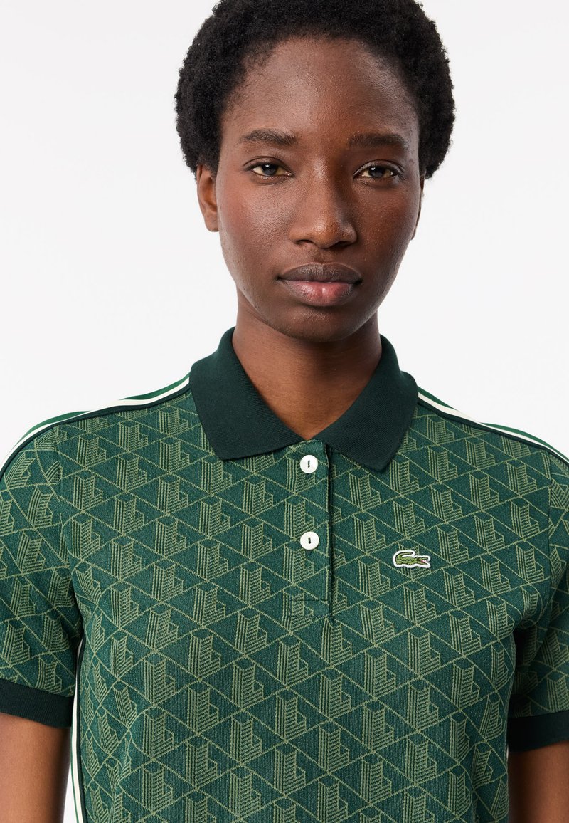 Zielona koszulka polo z geometrycznym wzorem, dwoma guzikami przy kołnierzyku i kontrastującymi ciemnozielonymi wykończeniami. Logo Lacoste na klatce piersiowej.