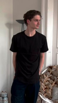 Zwarte t-shirt met een ronde hals, gecombineerd met donkerblauwe broek. De omgeving heeft een lichtgekleurde muur en decoratieve elementen op de achtergrond.