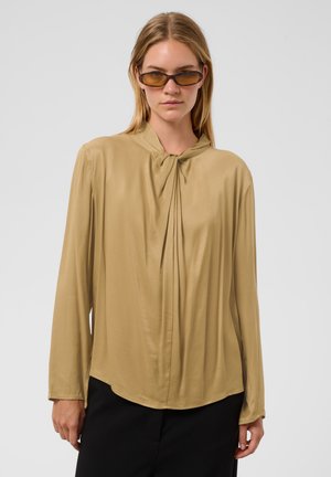 Mujer con blusa beige de manga larga con cuello plisado y gafas de sol con tintado ámbar, de pie frente a un fondo liso de color gris claro.