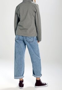 Veste grise à col, jean ample en denim bleu clair retroussé aux chevilles, et baskets montantes bordeaux. Tissu texturé avec coutures visibles.