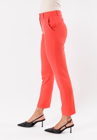Pantaloni tailoring corallo con vestibilità slim, fronte piatto e tasche laterali. Indossati con tacchi slingback neri con punta a punta.