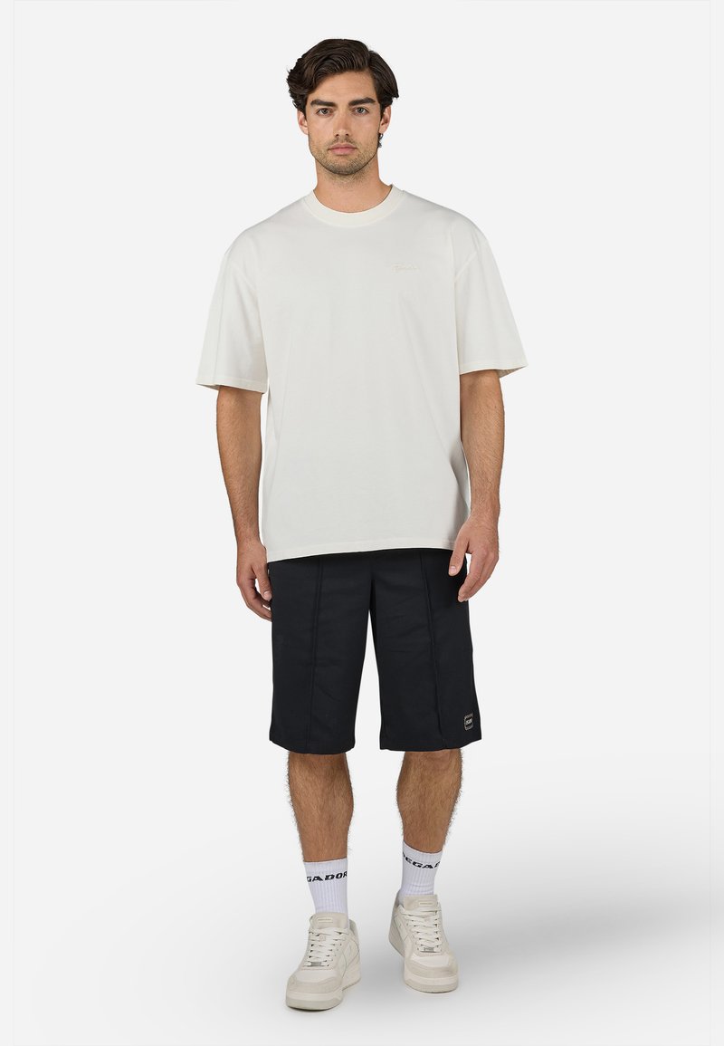 Kurzärmliges, rundhalsiges weißes T-Shirt kombiniert mit knielangen schwarzen Shorts, die ein dezentes Logo tragen, und weißen Sneakers mit strukturierten Sohlen.