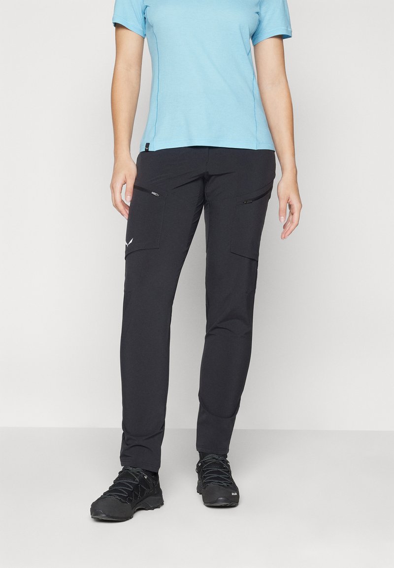 Calças pretas com bolsos de carga e fechos zíper, feitas de um tecido leve. Combinadas com uma camisa azul de manga curta e sapatos de caminhada pretos.