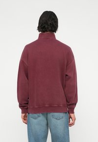 Sweatshirt bourgogne avec un col haut, coupe oversize, épaules tombantes et poignets côtelés. Porté avec un denim bleu clair.