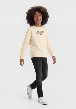 Nasmejani dječak hoda, nosi kremastu majicu s kapuljačom s logotipom "Jack & Jones", crne traperice i bijele tenisice, na jednostavnoj bijeloj pozadini.