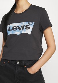 Šedé bavlněné tričko s modro-bílou grafikou mraku a textem "Levi's". Krátké rukávy a volný střih, doplněné tmavými džíny.