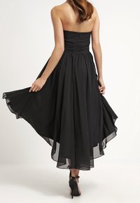 Femme en robe noire sans bretelles avec un ourlet asymétrique fluide, tenant les bords de sa jupe, portant des talons hauts noirs, photographiée de dos.