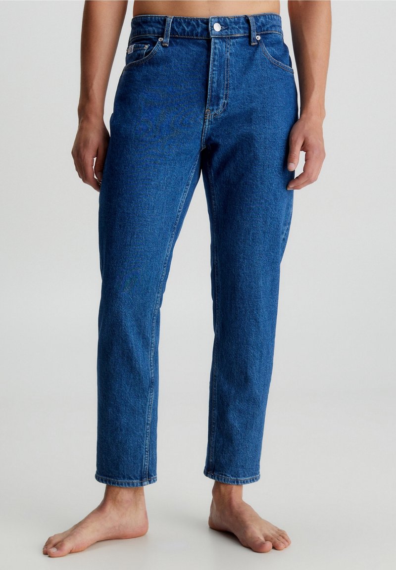 Mann trägt hochgeschnittene blaue Jeans, steht barfuß vor einem schlichten hellen Hintergrund, die Hände locker an den Seiten.