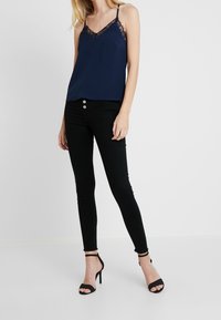 Camisole en soie bleu marine avec des bordures en dentelle noire, assortie à un jean skinny noir avec des boutons métalliques et des talons à lanières.
