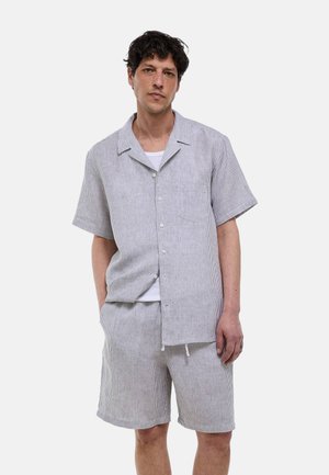 Homme portant une chemise rayée grise et blanche à manches courtes avec boutons et un short assorti jusqu'aux genoux, debout les mains dans les poches.