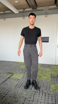 Młody mężczyzna stojący w pomieszczeniu, ubrany w czarną koszulkę, szare spodnie i czarne buty, na wzorzystej wykładzinie.