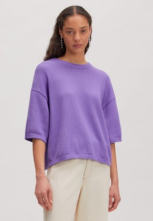 Femme portant un pull en maille lavande à manches courtes avec un col rond et un pantalon crème sur un fond uni.