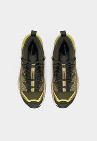 Ζευγάρι πράσινων και κίτρινων παπουτσιών trail running Salomon με μαύρα κορδόνια, σε λευκό φόντο, από ψηλά.