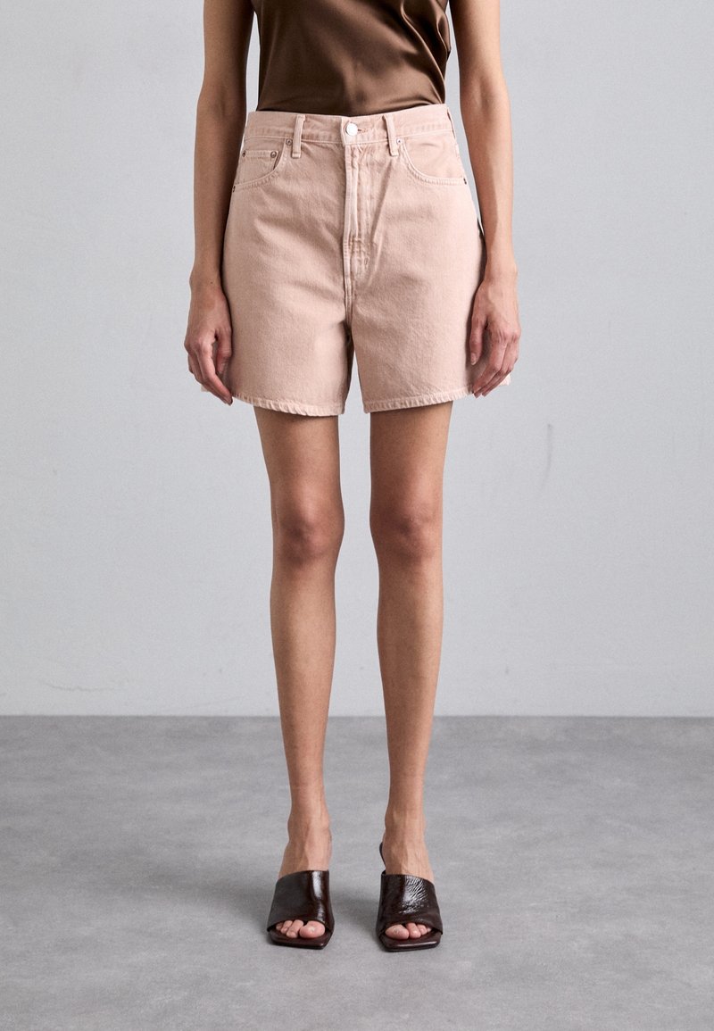 Pantalones cortos de mezclilla rosas claros con una cintura alta, cinco bolsillos y dobladillo sin terminar. Combinados con sandalias de tacón abierto en marrón oscuro.