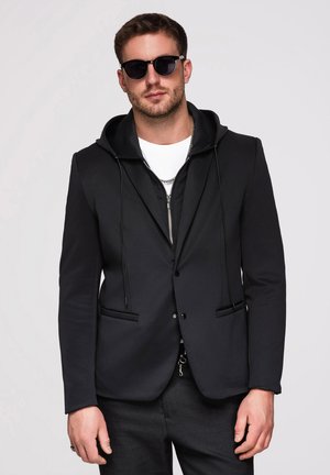 Homme portant un sweat à capuche zippé noir sous un blazer noir, une chemise blanche, des lunettes de soleil et un collier en chaîne argentée sur fond clair.
