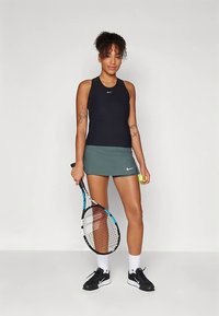 Camiseta sin mangas negra y falda de tenis verde; sosteniendo una raqueta de tenis y una pelota; exhibido sobre un fondo neutro. Logotipo de Nike en la ropa.