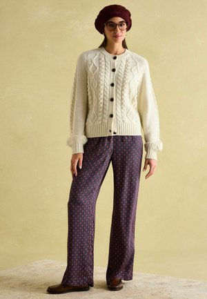 Cremiger Strickcardigan mit schwarzen Knöpfen, kombiniert mit navy-blauen, gemusterten weit geschnittenen Hosen mit roten Akzenten und einem burgunderfarbenen Barett. Braune Schuhe.