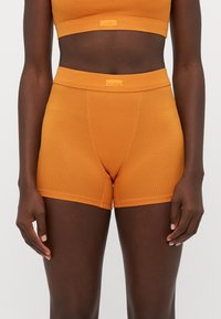 Persona di carnagione scura che indossa pantaloni corti a vita alta arancioni a coste e un crop top abbinato, in piedi contro uno sfondo chiaro uniforme.