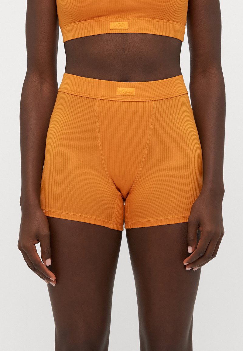 Persona di carnagione scura che indossa pantaloni corti a vita alta arancioni a coste e un crop top abbinato, in piedi contro uno sfondo chiaro uniforme.