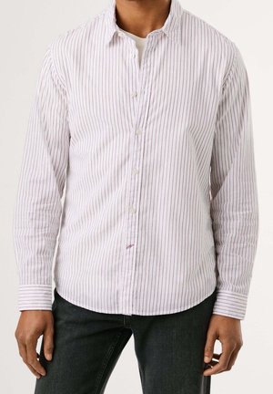 Hombre con camisa blanca abotonada con finas rayas rojas verticales y vaqueros negros, de pie sobre un fondo liso y claro.