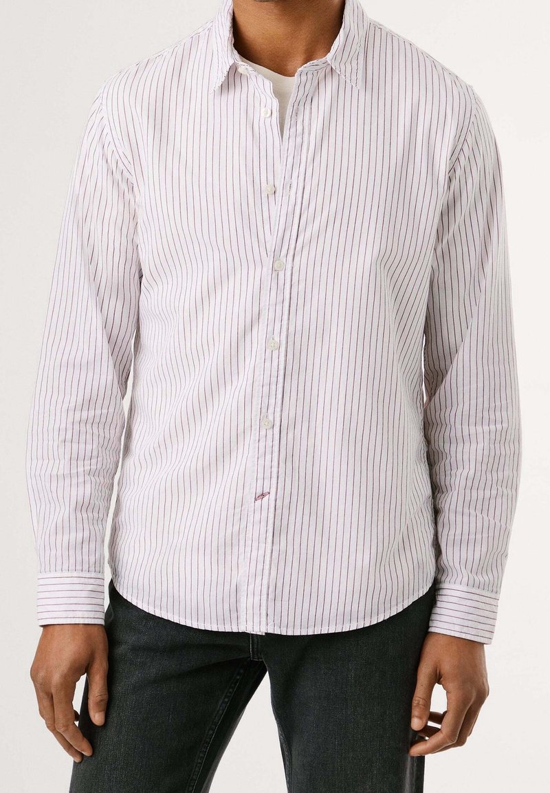 Homme portant une chemise blanche à boutons avec de fines rayures verticales rouges et un jean noir, debout devant un fond uni et clair.