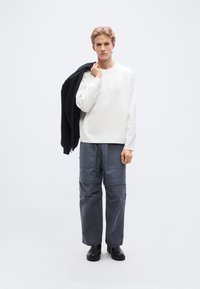 Λευκό πουλόβερ, γκρι φαρδιά παντελόνια, και μαύρα υποδήματα. Το μοντέλο κρατάει μαύρο μπουφάν στον ώμο. Λείο ύφασμα, χαλαρή εφαρμογή, μινιμαλιστικός σχεδιασμός.