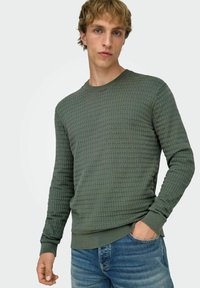 Jeune homme aux cheveux châtain clair portant une chemise texturée à manches longues de couleur vert olive et un jean bleu, une main dans la poche.