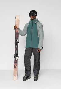 Férfi sídzsekit, nadrágot, csizmát és szemüveget viselve, egy FS márkájú sípárt tartva, függőlegesen egy sima világos háttér előtt.