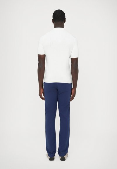 Biele rebrované tričko s krátkym rukávom v kombinácii s modrými nohavicami, zobrazené zozadu. Tričko má plochý golier a precízne detaily švov.