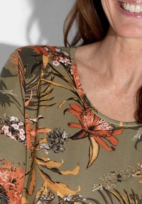 Bloemige top met korte mouwen, gemaakt van zacht materiaal. Het heeft een groene basis met oranje, gele en witte bloemenpatronen en een ronde halslijn.