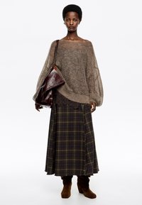 Žena oblečená vo voľnom hnedom svetri, kvetinovej dlhé sukni, hnedých čižmách a nesúca veľkú tmavohnedú koženú tašku na pleci.