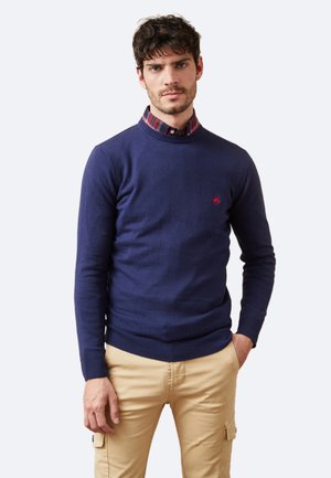 Suéter de punto navy con cuello redondo, puños y dobladillo acanalados, con un pequeño logotipo rojo. Llevado sobre una camisa de cuadros con cuello.