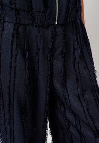 Navy textuur broek met verticale strepen en een rafelige afwerking, voorzien van een elastische tailleband. Inclusief een zichtbare zilveren ritsdetail.