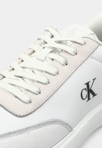 Biele tenisky vyrobené z semišu a kože, s mäkkým ružovým jazykom, bielymi šnúrkami a čiernym logom "cK" na boku.