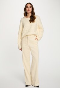 Donna in piedi con una mano in tasca, che indossa un maglione crema testurizzato e pantaloni a gamba larga in corduroy crema, con scarpe nere a punta.