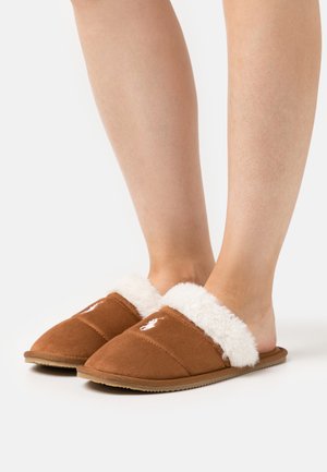 Chaussons en suède marron avec une doublure en fourrure blanche, semelle plate et détails cousus. Présente un logo sur le dessus.