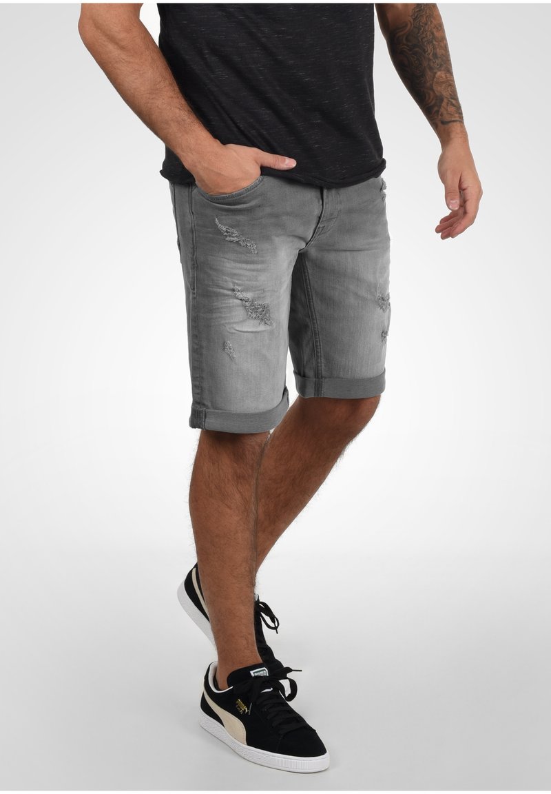 Shorts en denim gris avec des détails effilochés et des ourlets retroussés, associés à des baskets noires et dorées. Le tissu est légèrement usé et texturé.