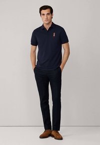 Marinblå pikétröja med korta ärmar, med ett broderat märke på bröstet. Kombinerad med mörka byxor och bruna snörskor.